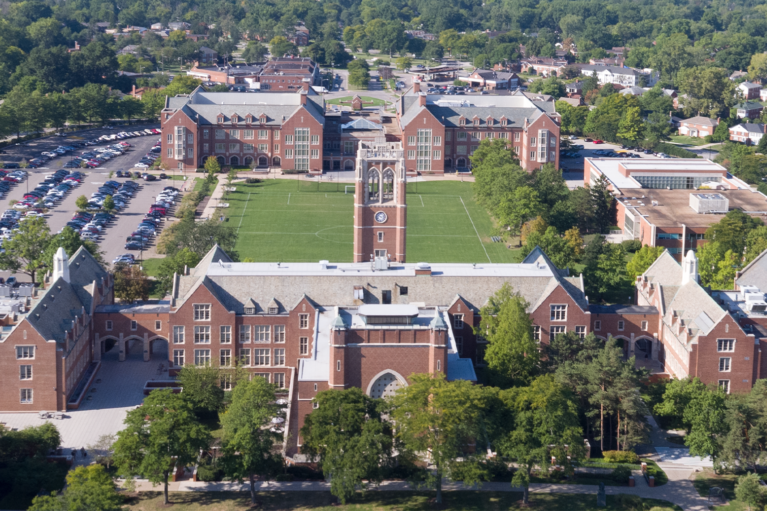 Aerial view of a college campus featuring red brick buildings, a clock tower, green athletic field, and parking lot.